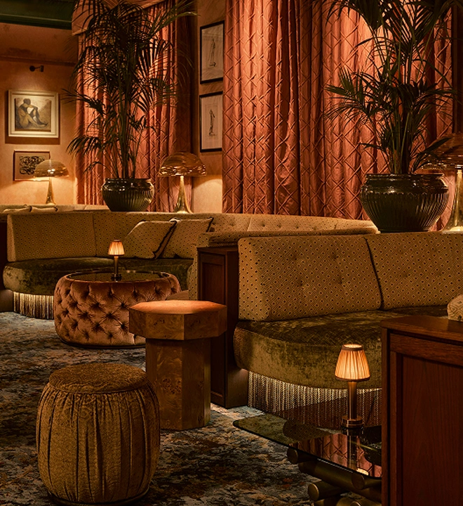 Cozy lounge with velvet sofas, round ottomans, warm lighting, plants, and framed art on curtains.
