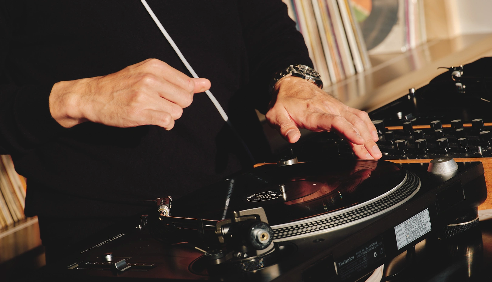Close-up of a DJ wearing a black long-sleeve shirt using a Technics turntable, one hand adjusting the vinyl record and the other holding the tonearm, with vinyl records on shelves in the background.