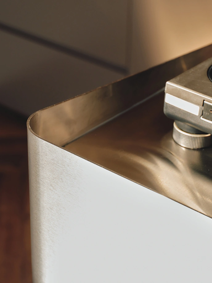 Close-up of a corner of a white and metallic gold table or cabinet with a rounded edge, featuring a portion of a vintage camera resting on top.