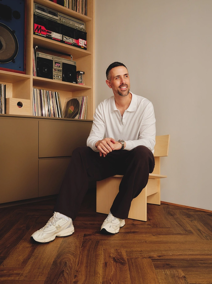 Man with short dark hair and beard sitting on a low wooden chair on wood floor, wearing a white quarter-zip sweater, black pants, white sneakers, and a watch, smiling and looking to the side. Behind him is a shelf filled with vinyl records, audio equipment, and speakers.