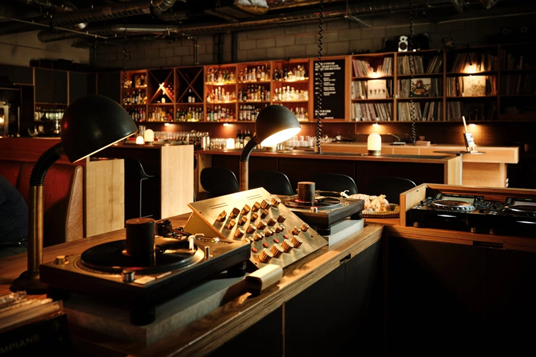Interior of a dimly lit bar with a wooden DJ booth featuring turntables, a mixer, and lamps, with shelves of bottles and records in the background.
