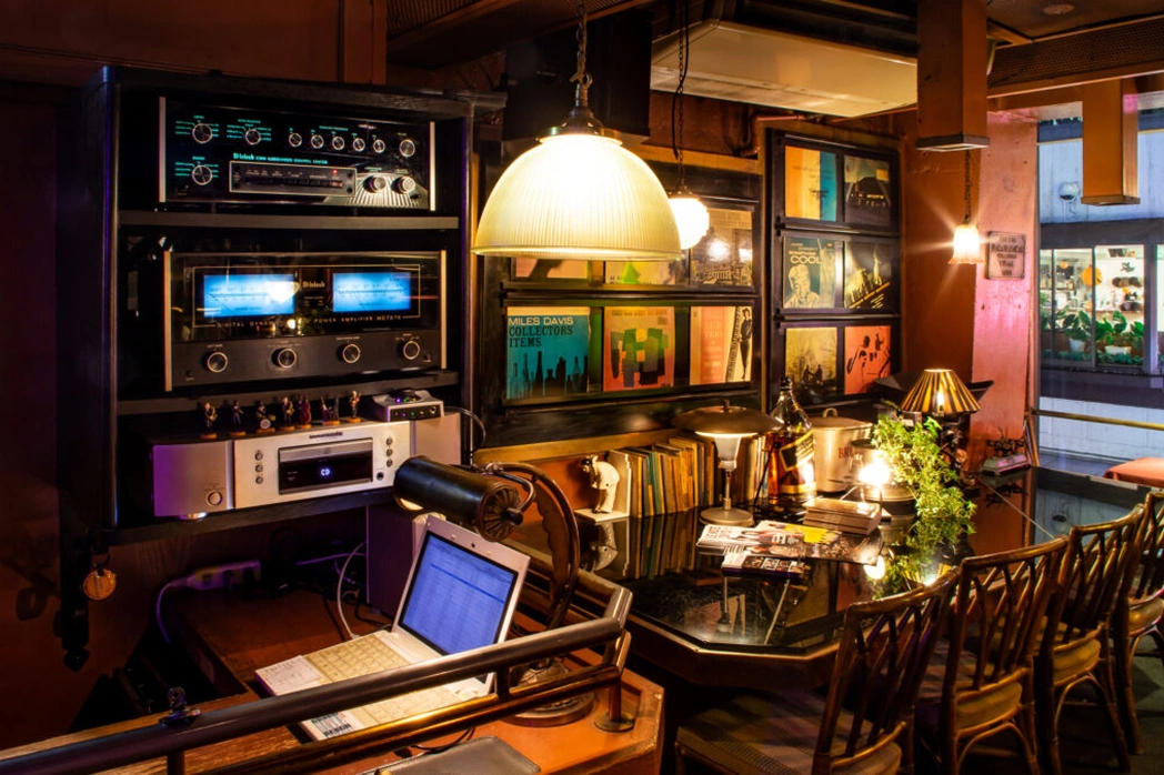 Cozy retro-style interior with a vintage music system mounted on the wall, including amplifiers and receivers. Below it, an open laptop sits on a wooden desk with a leather desk lamp. To the right, a glossy black table surrounded by wooden chairs holds scattered magazines, books, a green plant, and warm lighting from multiple lamps. The walls are decorated with framed vinyl record covers and warm, ambient lighting enhances the nostalgic atmosphere.