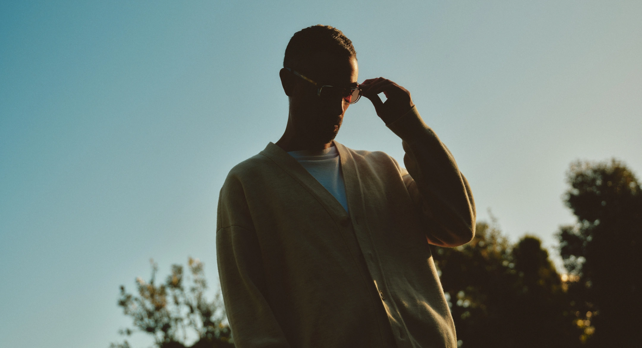 Silhouette of a person wearing glasses and a cardigan adjusting their glasses outdoors at sunset with trees in the background.
