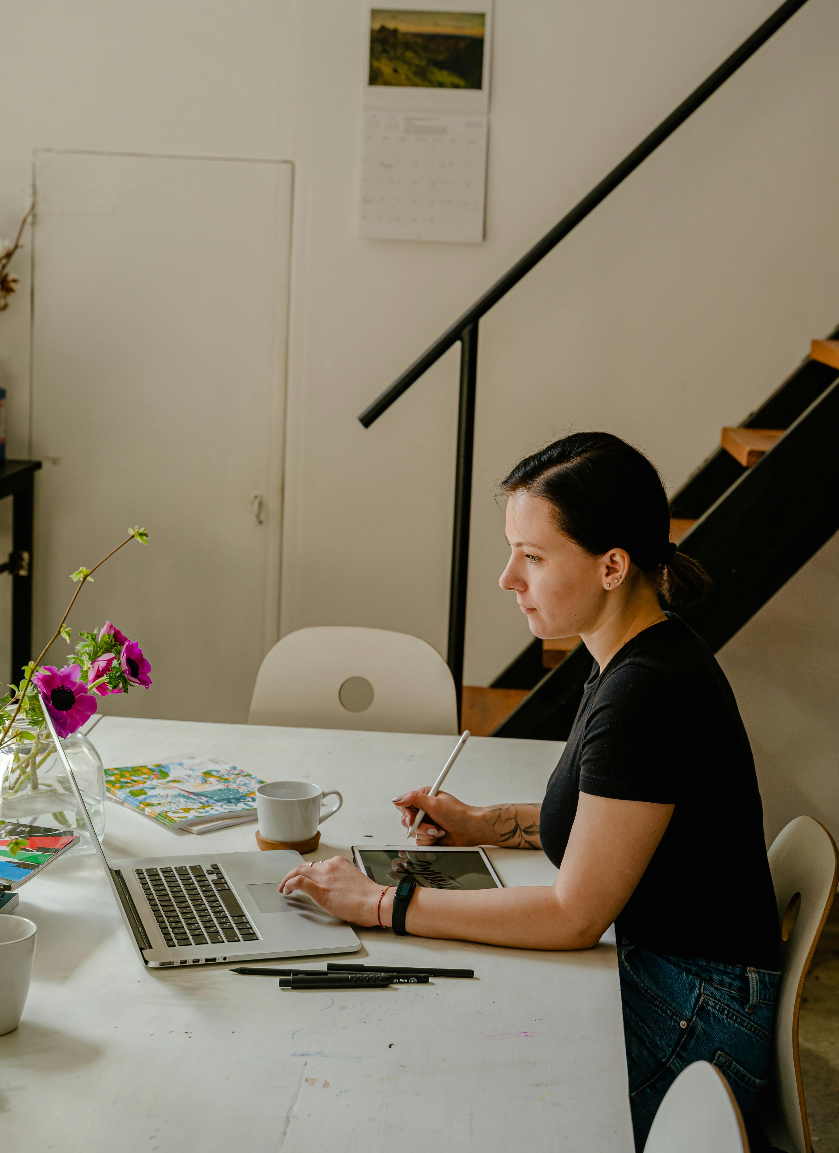 Woman working at a laptop at a white table with a notebook, a cup and a vase of flowers in a bright room