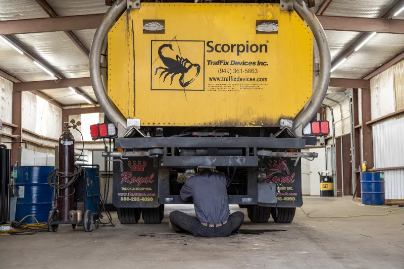 Mechanic working under a Scorpion crash truck with yellow TrafFix Devices panel in a service bay.