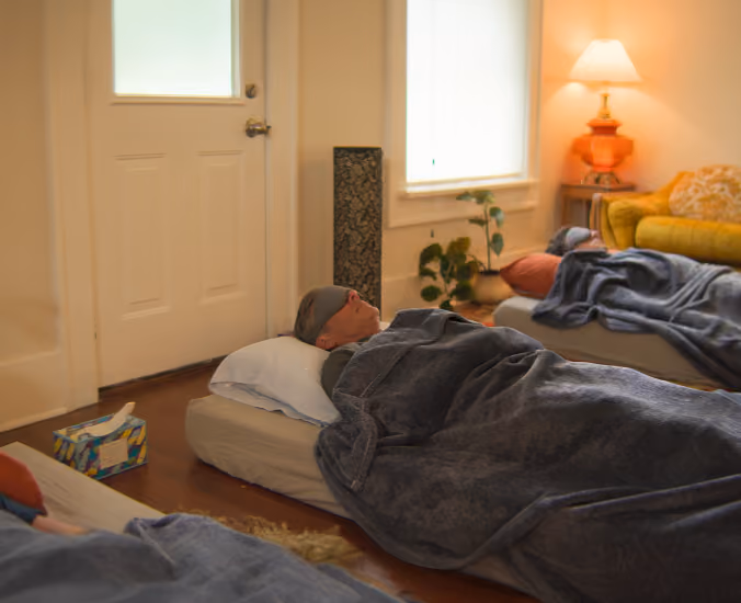 Person wearing a sleep mask lying on a mattress on the floor covered with a blanket in a cozy room.