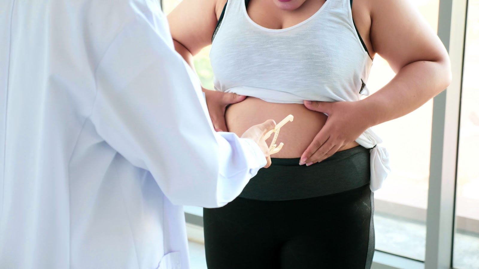 A female patient holds her belly fat while a doctor measures it during a medical evaluation.