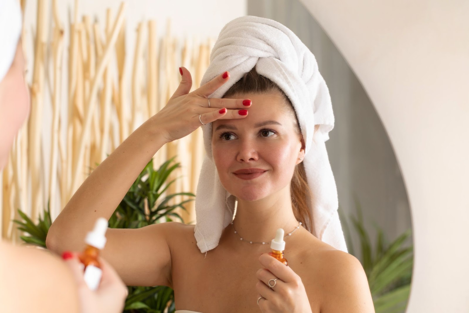 A woman applies facial serum on her face in front of a bathroom mirror.