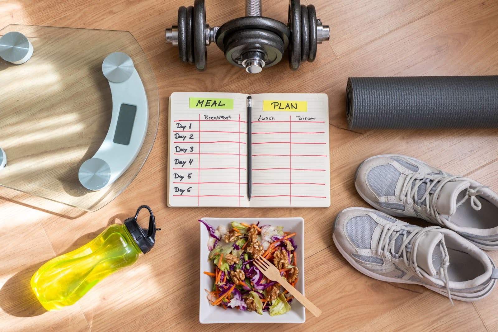 A meal planning notebook surrounded by a weighing scale, rolled exercise mat, running shoes, dumbbells, and a water bottle.