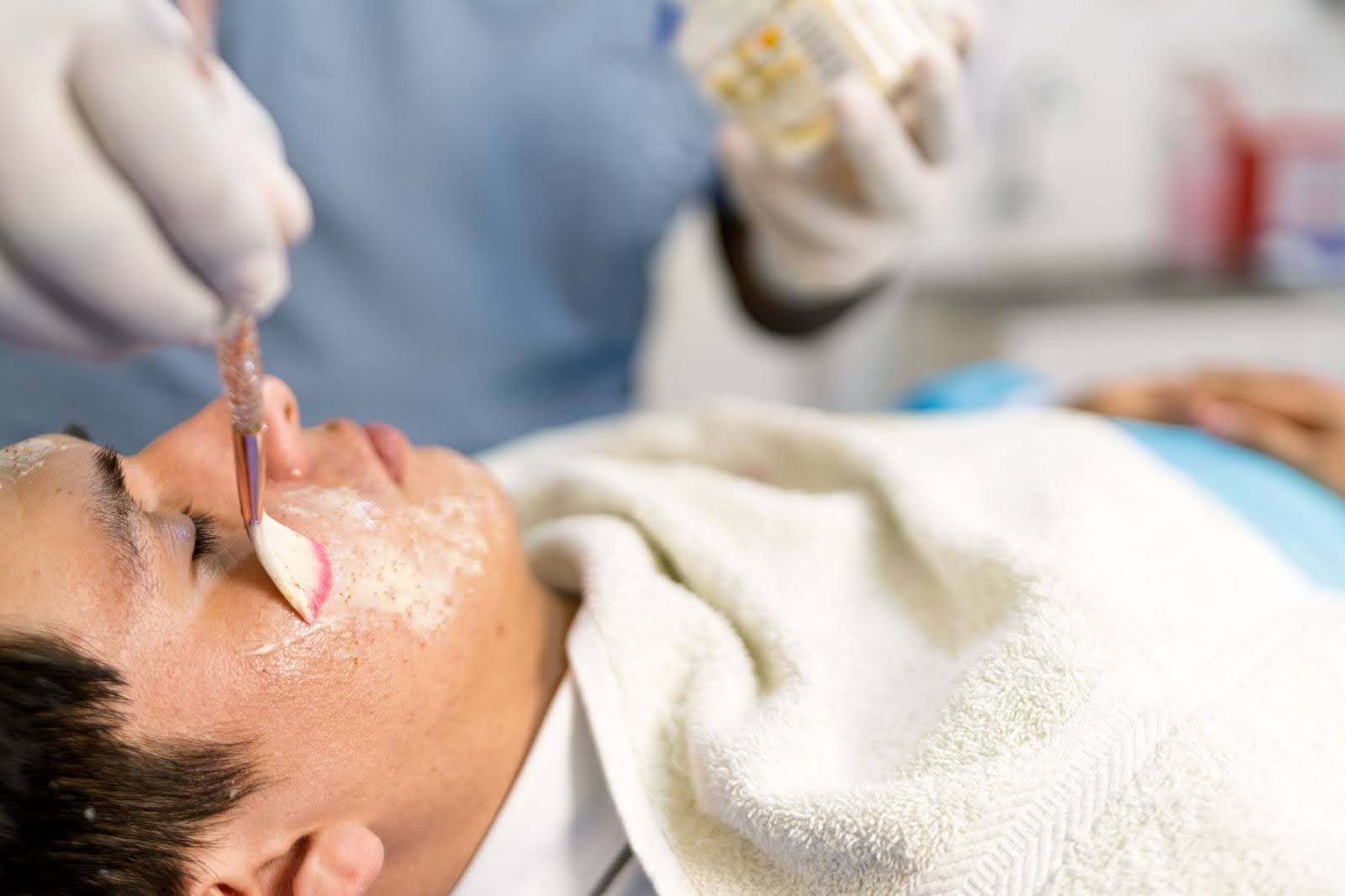 A skin professional applying an acne treatment mask during a facial on a young man’s face.