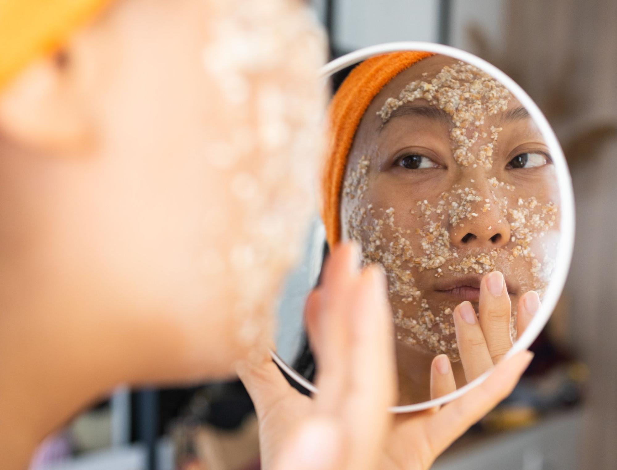 Asian woman gently exfoliating her face with oats while looking into a handheld mirror.