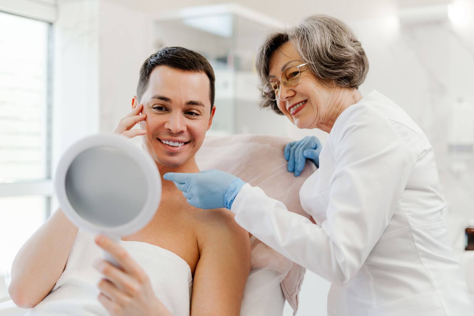 A male patient holding a mirror and discussing skin concerns with an experienced skin specialist.
