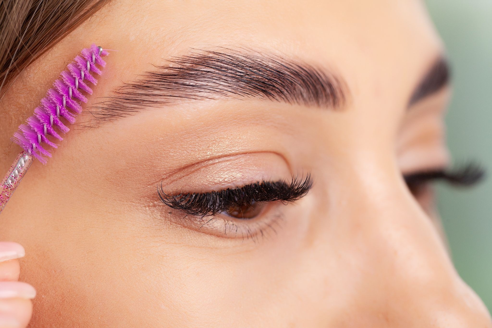 Close-up of a woman shaping and grooming her newly laminated brows using a pink brush