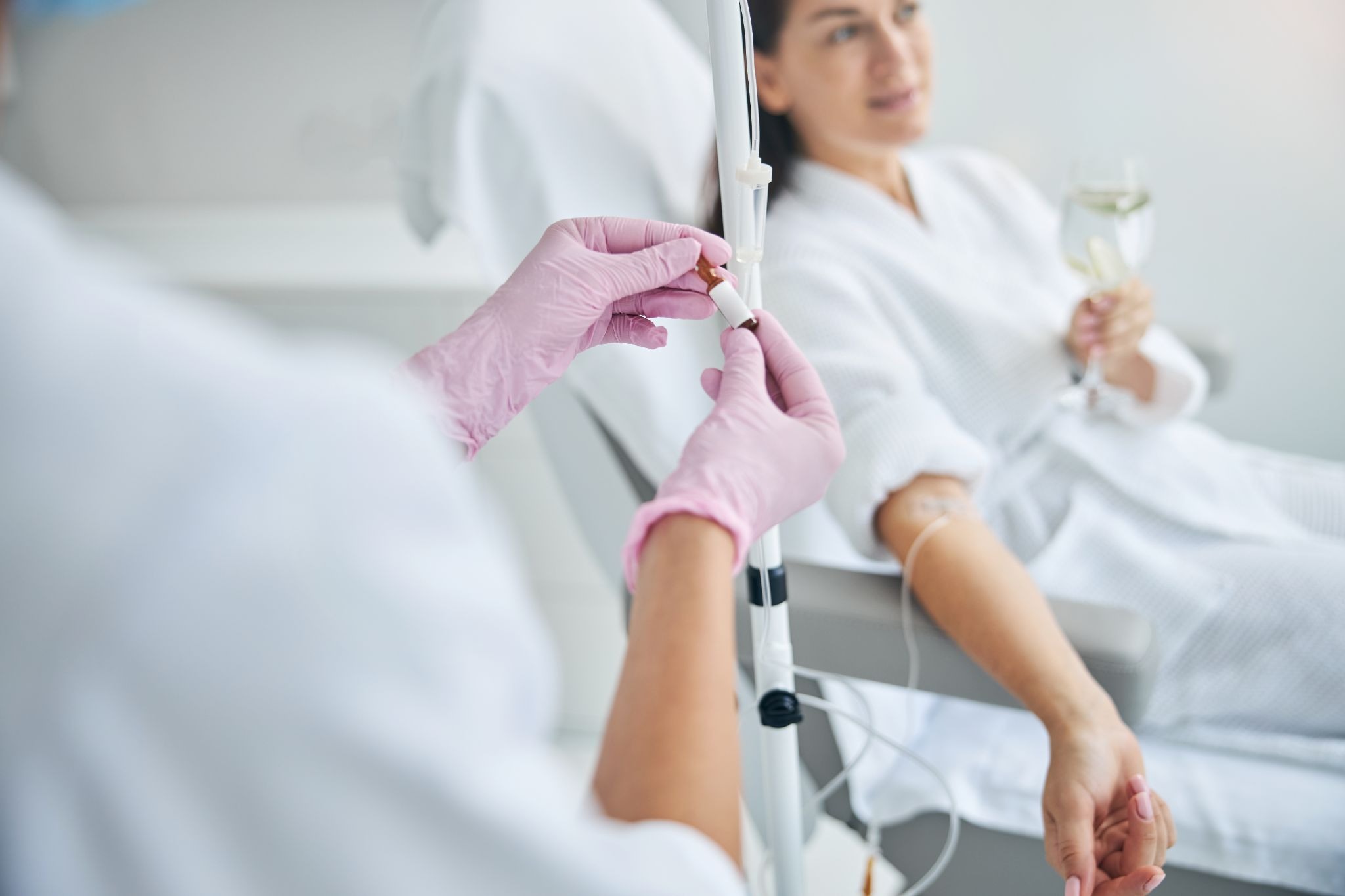 A nurse prepares a Vitamin IV therapy infusion for a female client, who is seated and holding a glass of water in the background