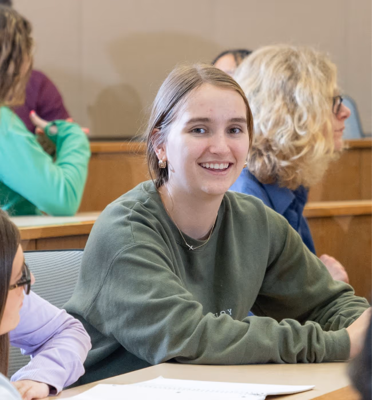 Student wearing a green sweatshirt sitting in a classroom and smiling.