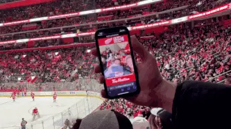 A group of smiling people live stream from a cell phone camera to a videoboard at a hockey arena.