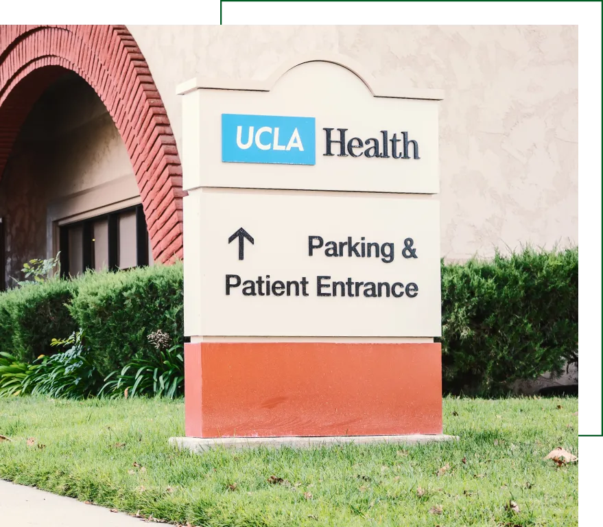 UCLA Health directional sign for parking and patient entrance outside healthcare facility with red brick arch in background
