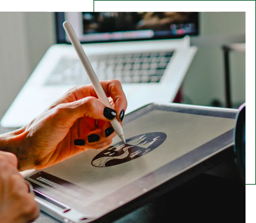 Close-up of person drawing a graphic on a tablet with stylus, laptop in background