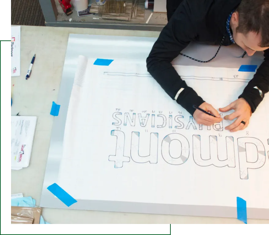 Designer outlining reversed lettering on a large signage template for 'Dmont PHYSICIANS' at a workspace table