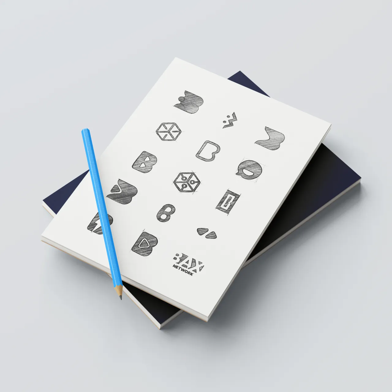 Niebieska kredka na białym szkicowniku z czarno-białymi szkicami logo i symboli, leżącym na dwóch ciemnych książkach.