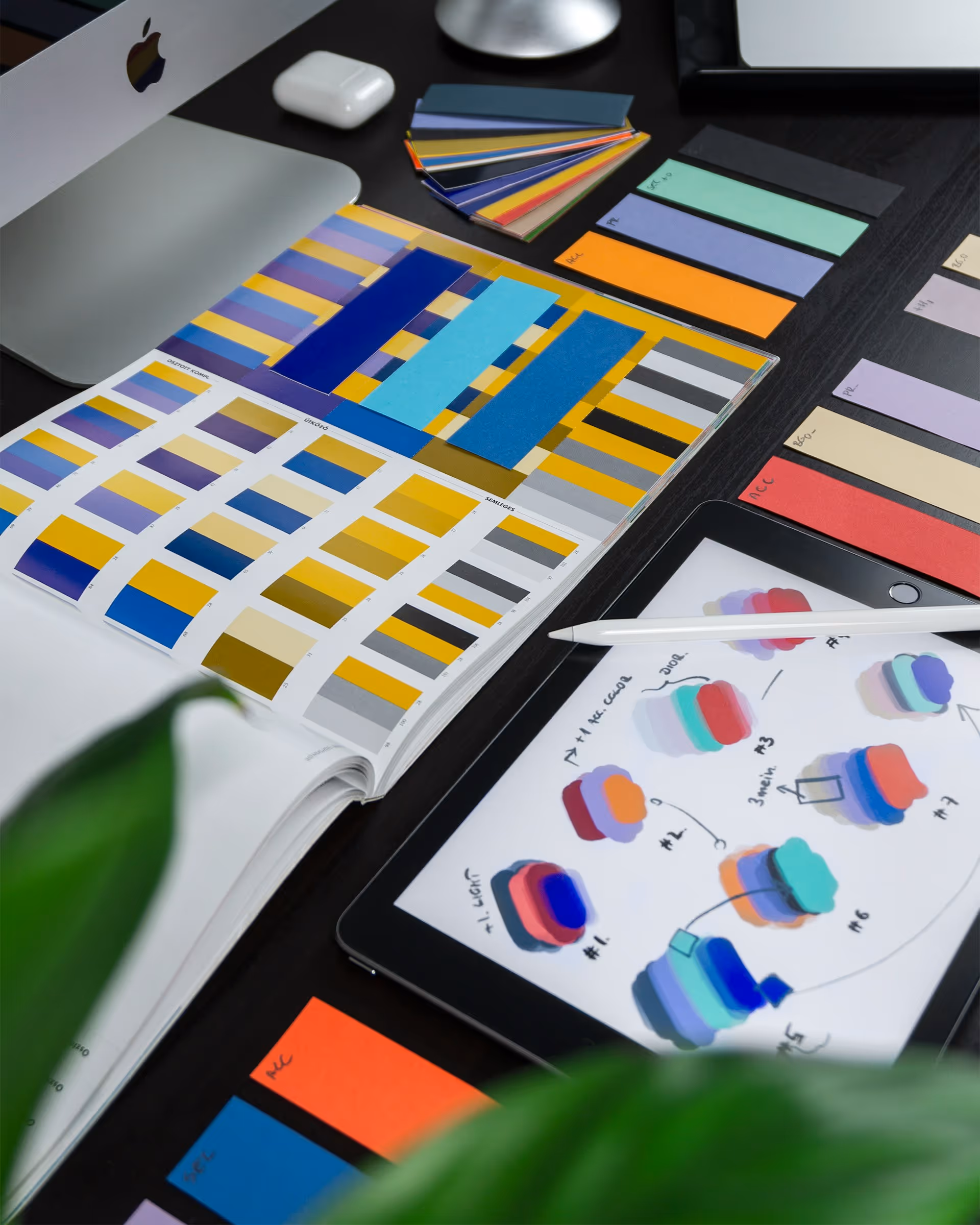 Stół z różnorodnymi wzornikami kolorów, tabletem z paletą odcieni i rysikiem oraz laptopem Apple w rogu.