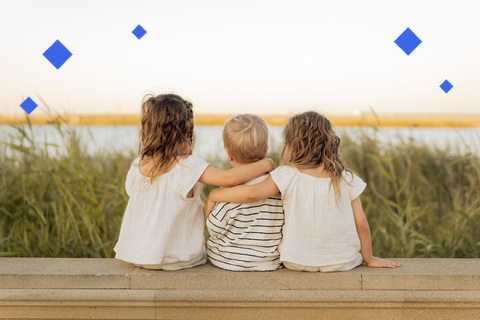 Three young siblings sitting together on a wall at sunset, arms around each other, seen from behind
