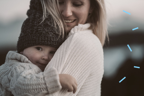 A mother in a knit sweater holding her young child close in a calm, tender outdoor moment