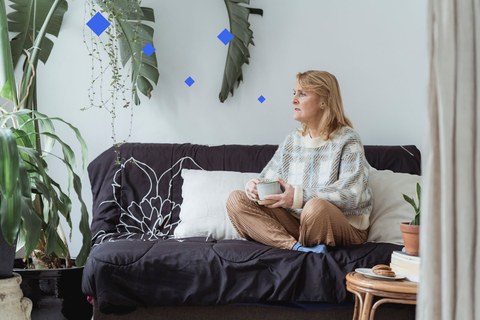 Woman in her fifties sitting on a sofa with a mug, pausing mid-thought and looking away