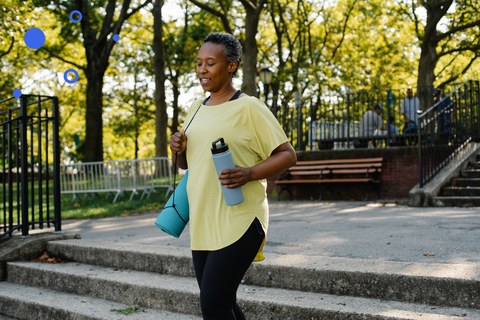 Woman in her fifties walking outdoors in a park with a water bottle and yoga mat