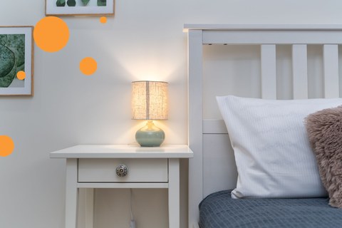Bedside table with a lit lamp beside a made bed in warm evening light
