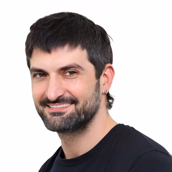Porträt eines lächelnden Mannes mit dunklen Haaren, Bart und schwarzem T-Shirt vor weißem Hintergrund.
