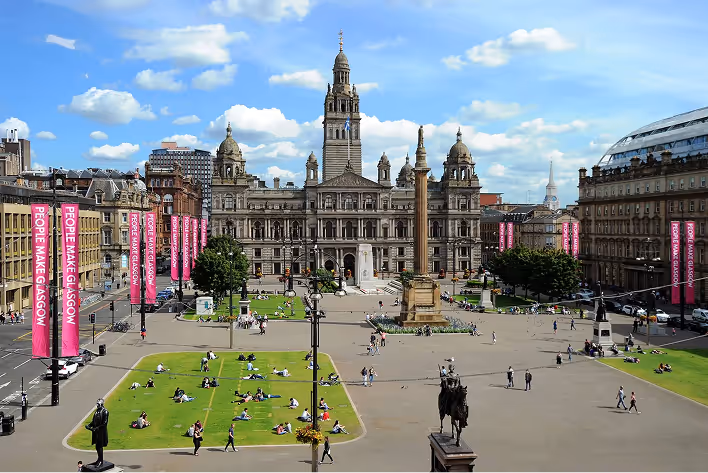 Glasgow city square with people on grass and historic city hall building under a blue sky.