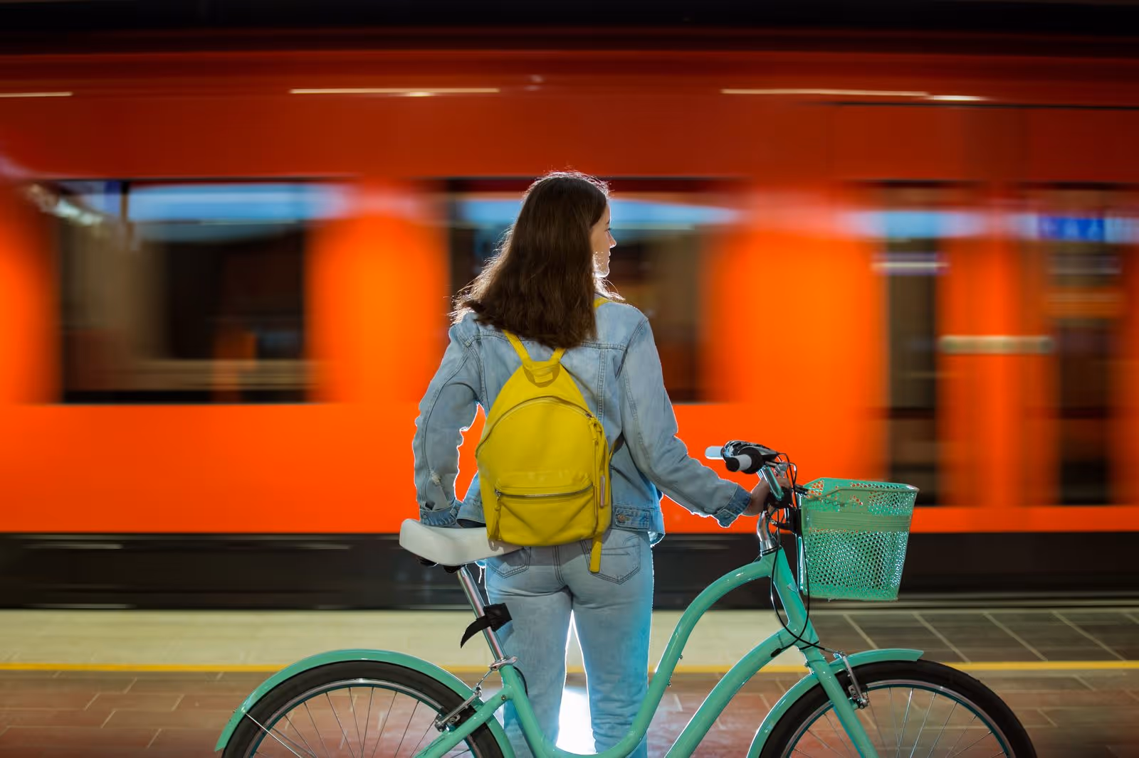 Woman with yellow backpack holding a teal bicycle on a subway platform as a red train passes by.