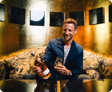 Smiling man seated on a patterned sofa pouring whiskey from a bottle into a glass in a warmly lit room with gold-toned walls.