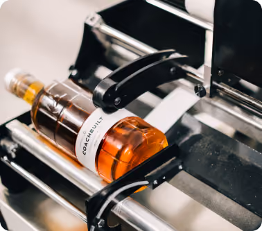 Bottle of amber liquid with a label reading 'COACHBUILT' being labeled by a machine.