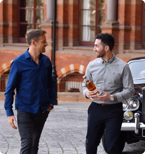 Two men smiling and walking outdoors near a vintage car, one holding a bottle of whiskey.