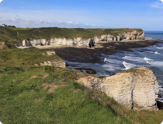Coastal landscape with white cliffs, grassy hills, and waves crashing on rocky shore under a blue sky.