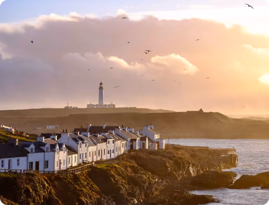 Coastal village with white houses on rocky cliffs and a lighthouse in the distance under a cloudy sky at sunset with birds flying.