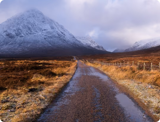 Wet country road leading towards snow-covered mountains under cloudy sky.