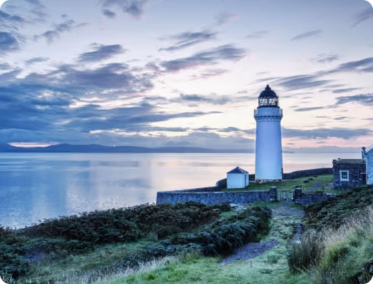 White lighthouse on a grassy coastline overlooking calm water under a partly cloudy sky at sunset.