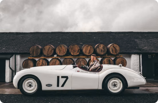 Man sitting in a vintage white racing car with number 17, parked in front of stacked wooden barrels.
