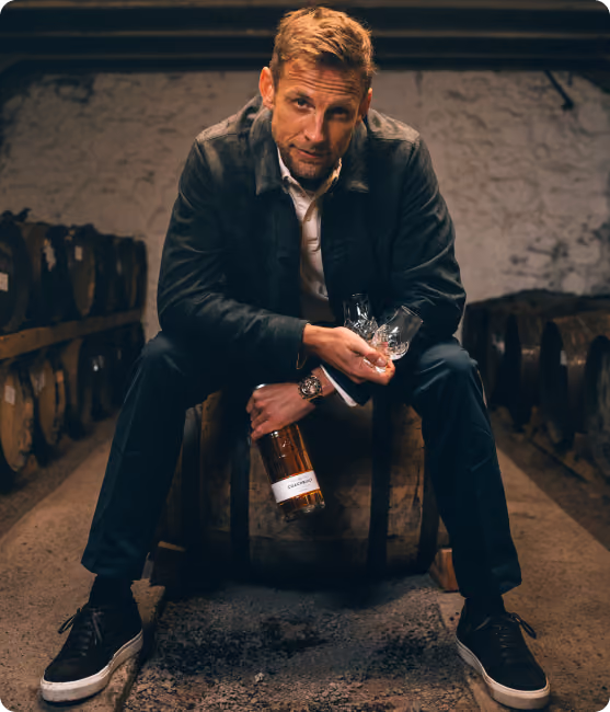 Man sitting on a wooden barrel in a cellar holding a bottle of whiskey and two glasses.