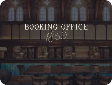 Interior of a vintage booking office with arched windows, wooden furniture, and a sign reading 'Booking Office 1869'.
