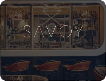 Interior view of a bar with shelves stocked with bottles and three brown bar stools, overlaid with the word 'SAVOY'.