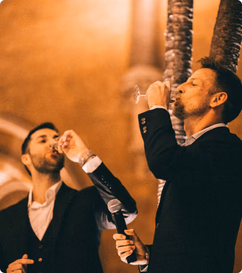 Two men in suits drinking from glasses, one holding a microphone indoors with warm lighting.