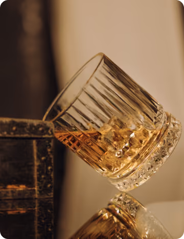 Glass of whiskey with ice cubes tilted on its side on a reflective surface next to a wooden box.