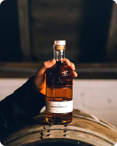 Hand holding a bottle of amber-colored Coachbuilt whiskey above a wooden barrel in a dimly lit setting.