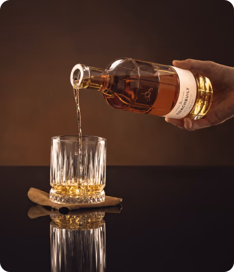 Hand pouring amber whisky from a bottle labeled 'The Sacherlilt' into a crystal glass resting on a cinnamon stick on a reflective black surface.