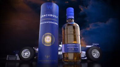Bottle of Coachbuilt Williams Racing 18-Year-Old whisky alongside its cylindrical blue packaging, set against a dark dramatic background with racing car tires.