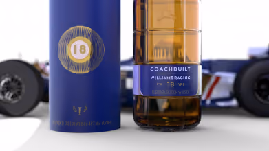 Close-up of a blue cylindrical box and bottle of Coachbuilt Williams Racing 18-year-old blended Scotch whisky on a white surface.